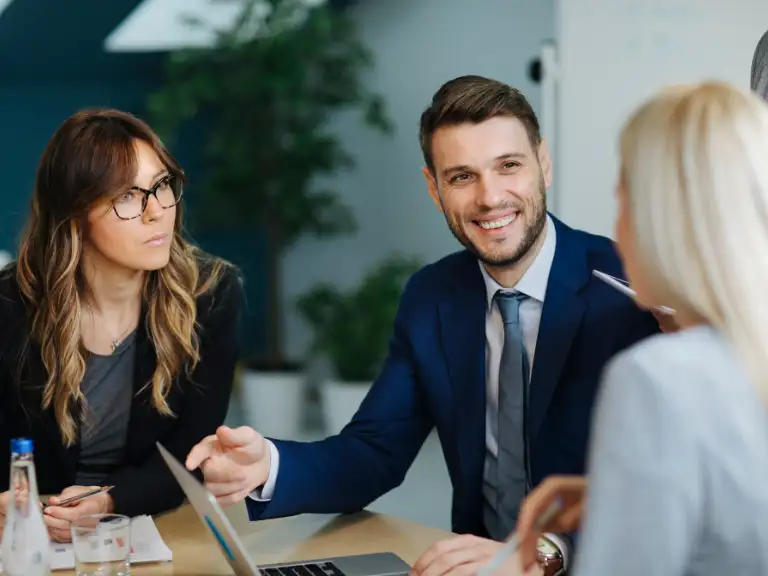 Profissionais em reunião, com um homem de terno azul sorrindo e gesticulando em direção a uma mulher loira, enquanto outra mulher de óculos observa, transmitindo interação e colaboração